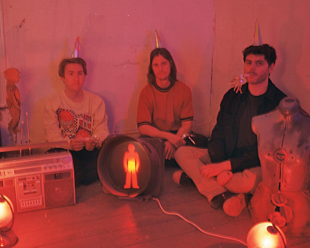 The three members of Eyesore & the Jinx sitting next to each other on the floor. They're wearing small conical party hats, and one has a party blower in his mouth. None of them are smiling.