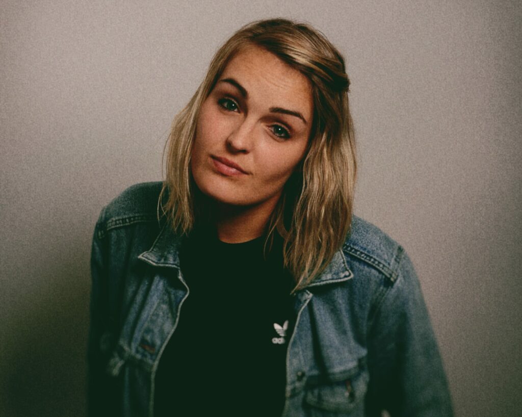 Informal, portrait style shot if Jen, a young woman with shoulder length blonde hair, Jen is wearing a black t-shirt with a white Adidas logo underneath a blue denim jacket