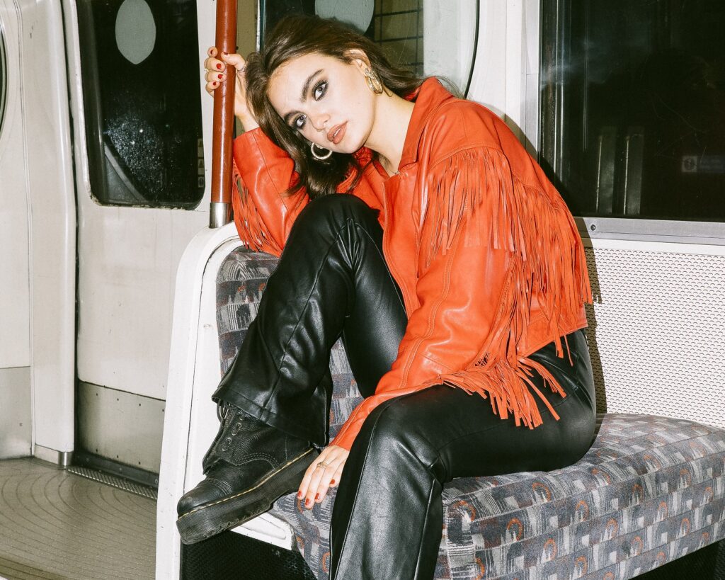 Viia, a young woman wearing a fringed orange jacket, black trousers and black chunky boots, sits on the seat of a train