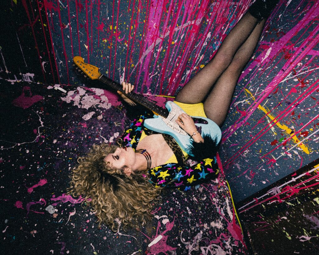 Cortney Dixon sits in a paint filled room playing guitar