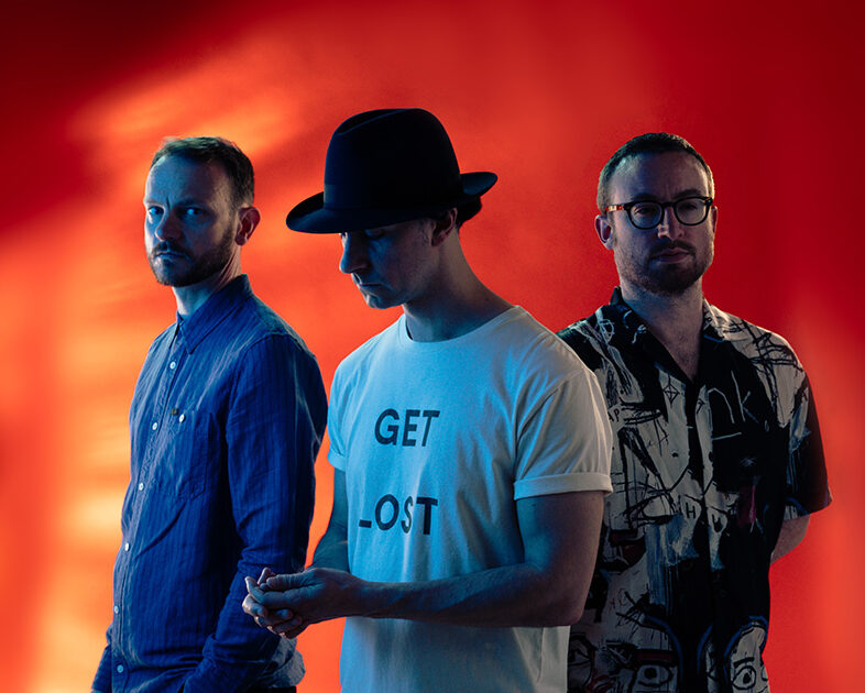 the three members of Maximo Park standing in a row against a dramatic red-orange gradient background. From left to right: a member wearing a blue button-up shirt, the middle member wearing a black fedora hat and white t-shirt with "GET LOST" text, and a member wearing glasses and a black and white patterned shirt. The lighting creates a moody, atmospheric effect with sharp shadows and highlights. The image has a professional, editorial quality with careful composition and stylized lighting.